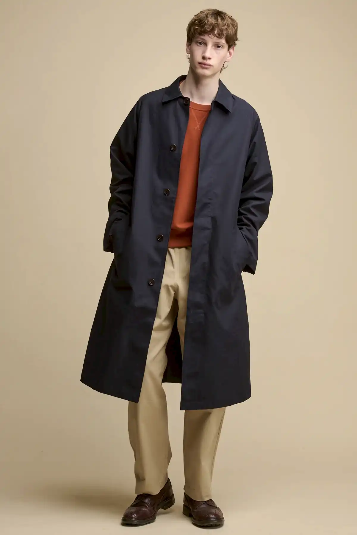 Full body image of the front of male wearing unfastened Frank raglan belted Raincoat in navy worn over raglan sweatshirt in cinnamon and cameraman pants in putty 