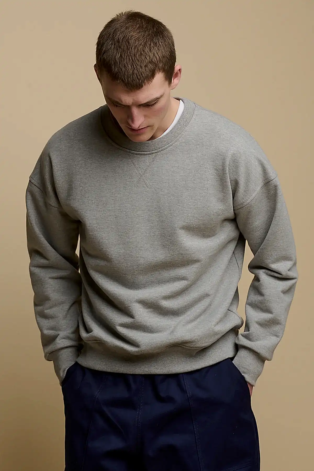 Thigh up front of brunet male looking down to the ground, wearing men's heritage drop shoulder sweatshirt in grey, worn over crew neck white t shirt paired with cameraman pants in navy