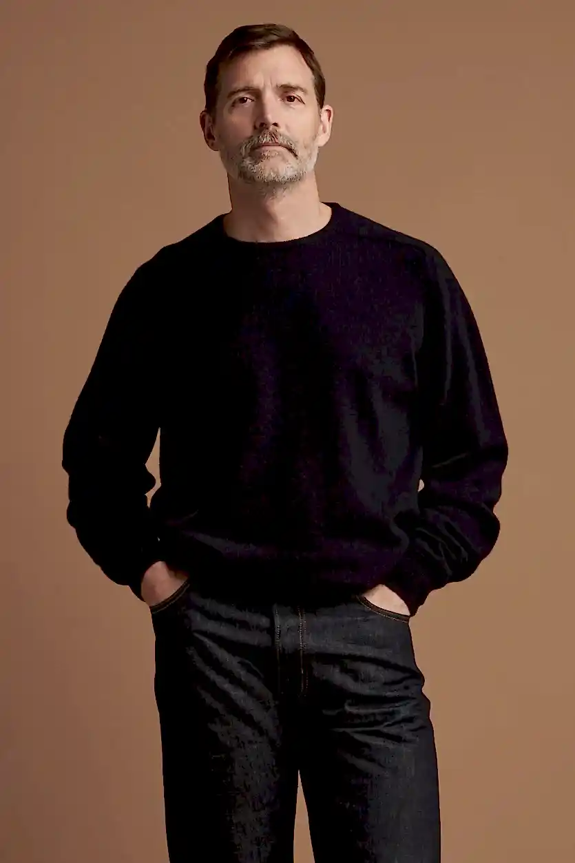 Thigh up front of Patrick Grant wearing lambswool saddle shoulder crew neck jumper in navy, worn with loose fit button fly Arran jeans in indigo